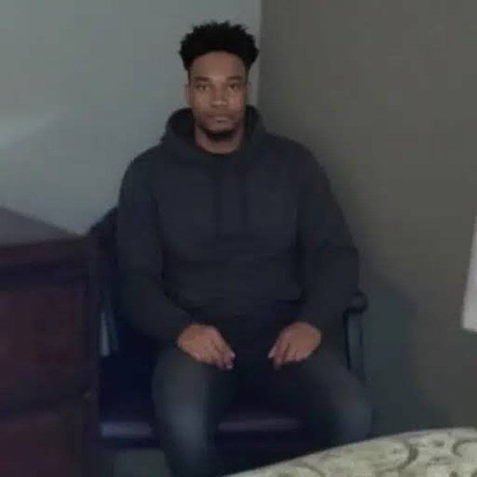 A man in a black hoodie sits motionless in a chair with a blank expression, humorously captioned as the imagined intense, serious posture the person will use while reading a tweet about alleged threats and hush money involving influencer $ANSEM.