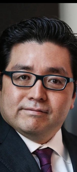 A close-up portrait of a middle-aged man with dark hair and glasses wearing a dark suit and purple tie.