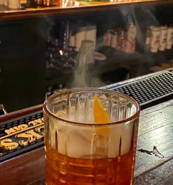 An Old Fashioned cocktail with a flaming orange peel garnish sits on a bar mat, casting a smoky haze against a backdrop of liquor bottles. The smoky presentation of the drink visually echoes the post's playful references to "smoking" and "smoke," while the dim bar atmosphere mirrors the moody, nighttime vibe of the caption.