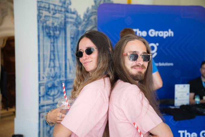 Two young people wearing matching pink shirts and sunglasses posing back-to-back in front of a blue 'The Graph' branded banner, holding iced drinks with red and white striped straws.