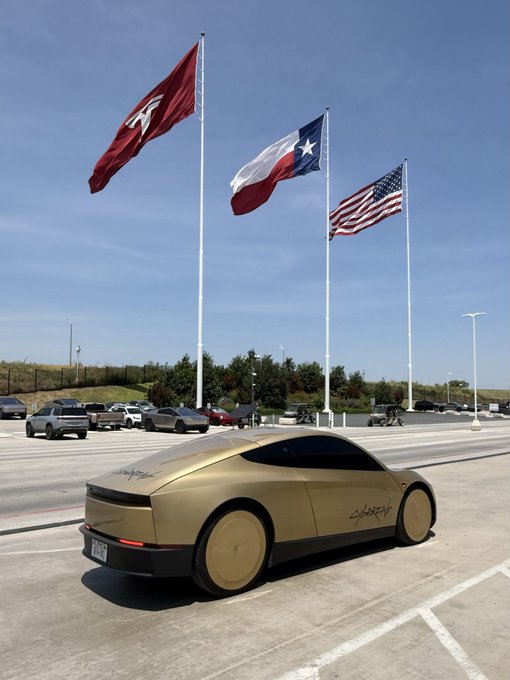 A matte gold Cybertruck prototype, bearing "CYBERTRUCK" lettering on its rear quarter panel, is parked in a lot beneath three flags flying against a clear blue sky—representing the Tesla logo, Texas, and the United States. The "GFTX" (likely a Texas license plate) and Texas state flag reinforce the location as Gigafactory Texas, where the Cybertruck is manufactured.