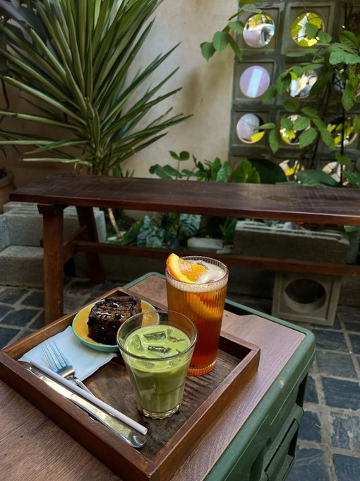 A wooden tray on a small table holds a green matcha drink with ice, a layered orange-colored beverage garnished with an orange slice, and a slice of chocolate cake, set in a garden courtyard.