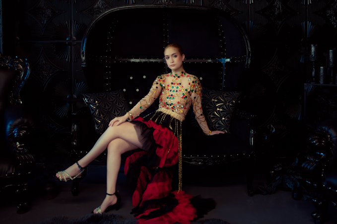 A fashion editorial photograph features a model seated on an ornate black couch wearing a sheer long-sleeved top adorned with colorful gemstones and a flowing red and black skirt.