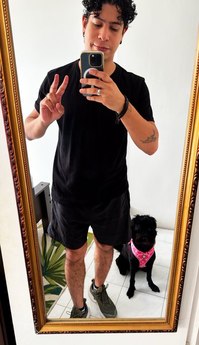 A mirror selfie of a young man in a black t-shirt and shorts making a peace sign, with a small black dog wearing a pink bandana sitting at his feet.