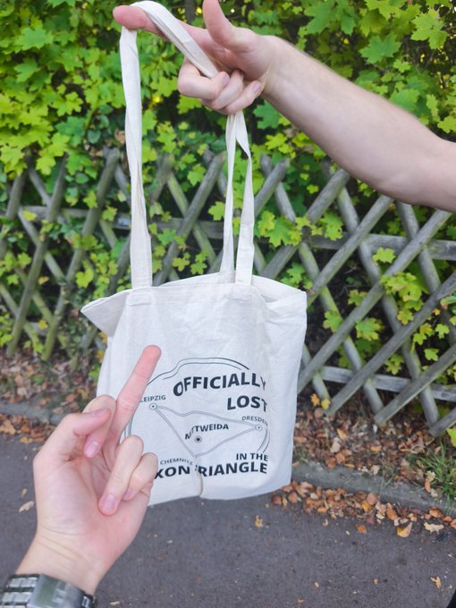 Two hands hold up a white tote bag printed with 'OFFICIALLY LOST IN THE BERMUDA TRIANGLE,' featuring locations LEIPZIG, DRESDEN, and CHEMNITZ, set against a green leafy background.