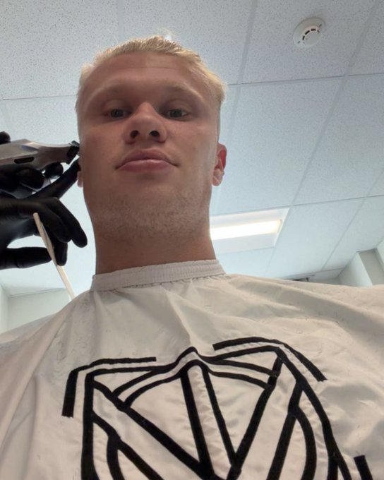 A young man with bleached blonde hair films a selfie video while a barber wearing black gloves prepares to cut his hair, with a black diamond-shaped graphic visible on his white shirt. The image relates to the post about Erling Haaland getting a fresh haircut before Norway's match against Brazil, referencing the narrative and historical betting odds mentioned.