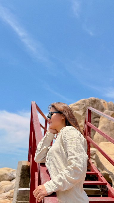 A woman in a beige hoodie and sunglasses stands on red stairs against a bright blue sky with wispy clouds, near rocky terrain.