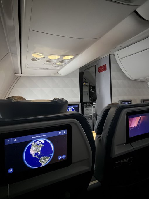 An empty airplane cabin interior is shown, featuring overhead compartments, lights, and seatback screens displaying a flight map and entertainment.