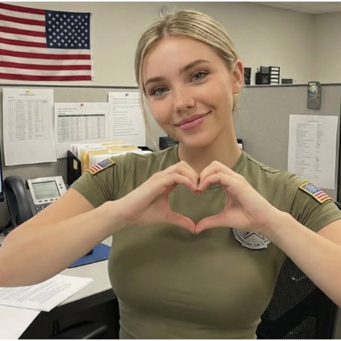 A young woman in an olive green t-shirt with American flag patches on the sleeves smiles and makes a heart shape with her hands in an office setting with an American flag in the background.