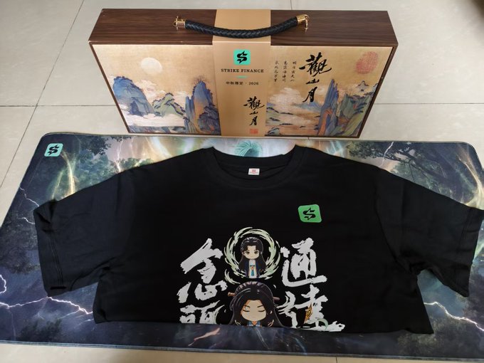 A black t-shirt with Chinese characters and character illustrations sits on a desk mat, with a decorative box labeled 'STRIKE FINANCE' and '中秋限定·2024' behind it.