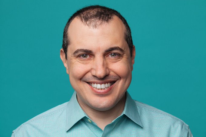 A smiling man with short dark hair and a light blue checkered shirt poses against a bright teal background, likely capturing the spirit of Andreas Antonopoulos, a respected figure in the cryptocurrency community whose wisdom and contributions are fondly remembered in the accompanying tribute.