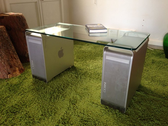 A custom desk made from a glass top and two silver Apple Power Mac G5 computer towers serving as legs, sitting on green carpet.