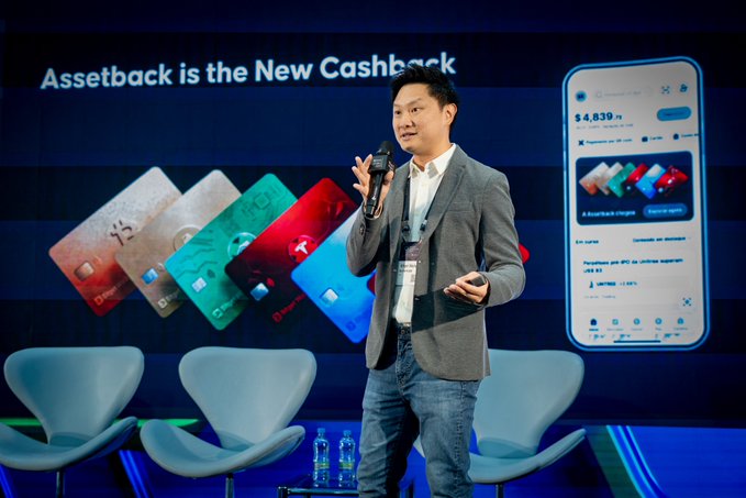 A speaker presents the "Assetback is the New Cashback" concept at the Blockchain Rio event, showcasing crypto debit cards and a digital wallet interface displaying a $4,839 balance with assetback rewards.