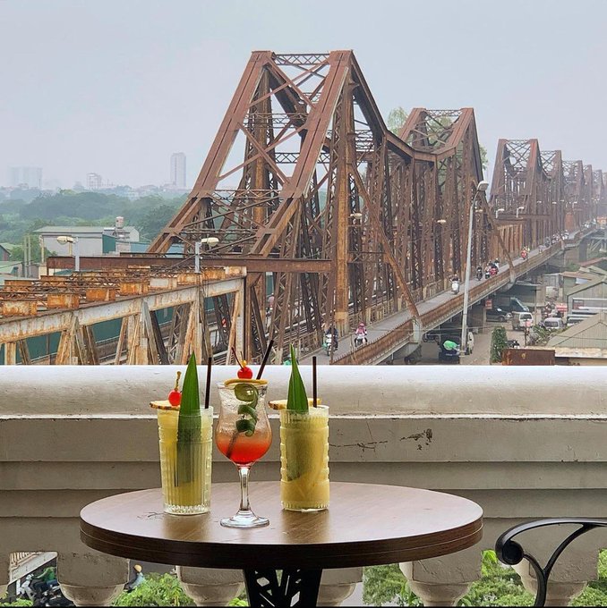 Three refreshing cocktails rest on a wooden table at a rooftop café, with the iconic rust-colored Long Biên Bridge and bustling Hanoi streets stretching out behind them, perfectly capturing the peaceful charm of the city that the post describes.