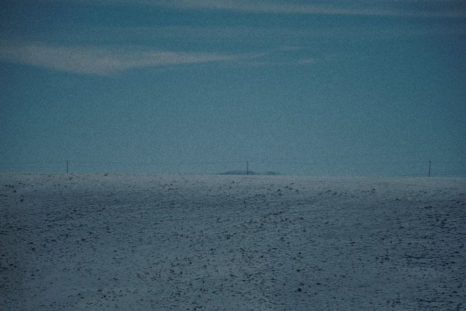 A minimal, muted landscape showing a vast, snow-covered plain under a gray-blue sky with faint clouds and distant utility poles on the horizon.