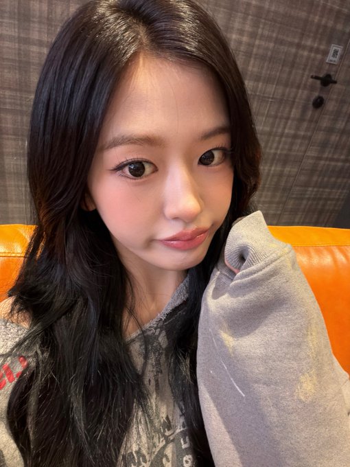 A close-up selfie of a young woman with long dark wavy hair, glossy pink lips, and dramatic eye makeup, wearing a distressed gray sweatshirt in front of an orange tufted leather seat.