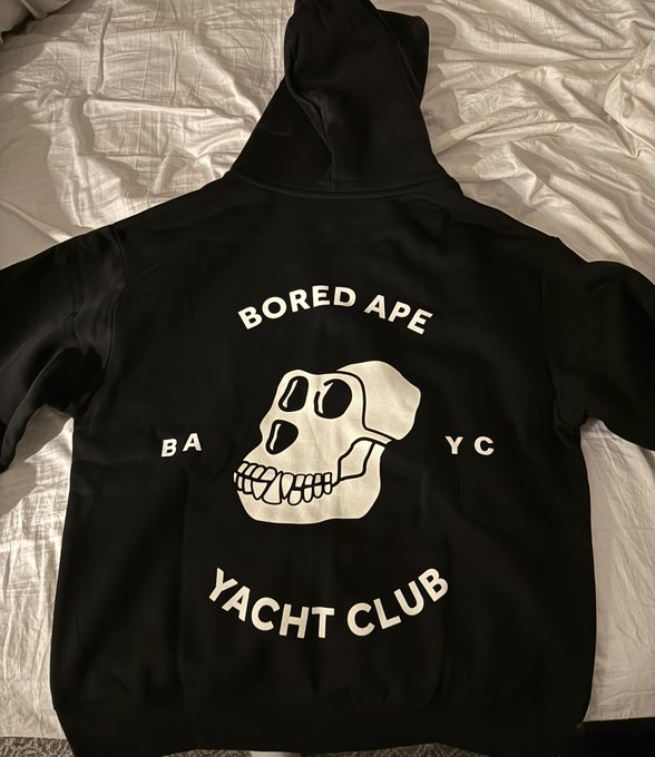 A black hoodie laid out on a white bed sheet, featuring the 'BORED APE YACHT CLUB' logo with the iconic skull graphic and 'BA YC' text on the sides.