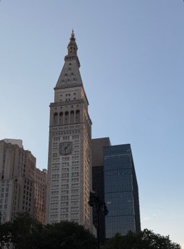 The image features a tall white building with a clock tower, which appears to be an old-fashioned structure. The sky above the cityscape is blue, creating a beautiful contrast between the building's architecture and the natural surroundings. In the background, there are other buildings that contribute to the urban environment.
