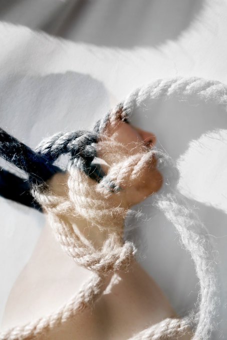 An artistic close-up of a hand bound with thick cream and dark blue rope, wrapped around the fingers against a bright white fabric background.
