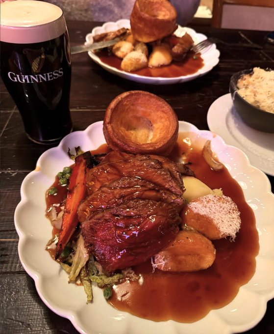 A traditional Sunday roast with roast beef, crispy potatoes, vegetables, and a golden Yorkshire pudding sits on a scalloped plate beside a pint of Guinness, capturing the perfect way to celebrate the day.