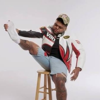 A man wearing a red, white, and black "Vans" racing-style jacket and a cap sits on a wooden stool, lifting his leg to pull off a white sock, with visible tattoos on his leg and arm. The image fits the caption's theme of playful disguise and subversion, visually echoing the idea of "infiltrating" a space under a casual, unassuming guise.
