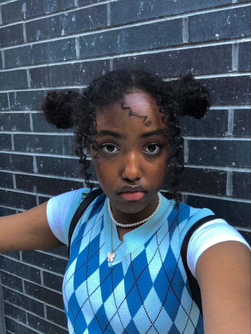 A young woman with curly hair styled in space buns, wearing a blue argyle sweater vest, posing in front of a dark brick wall.