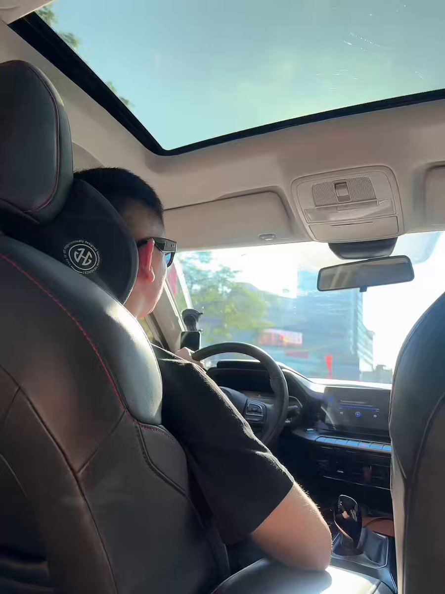 The image shows a man sitting inside a car with his back facing the camera. He appears to be wearing sunglasses and has a serious expression as he looks out of the window, possibly observing something interesting or worrying about traffic outside. The car's interior features black leather seats that match the man's attire.