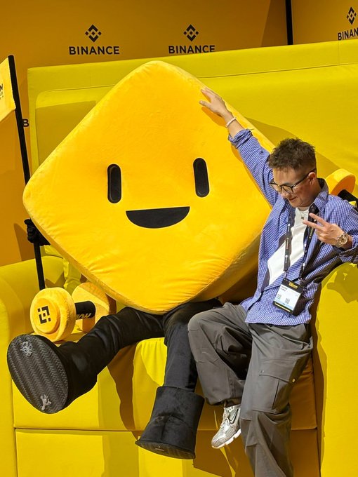 A person poses playfully with Bibi, the official Binance mascot plushie, at a Binance event booth featuring the prominent "BINANCE" branding in the background. The yellow square character with a smiling face, identified as $BIBI in the post, lounges alongside the attendee in a vibrant photo-op setting that captures the community-focused, fun spirit described in the caption.