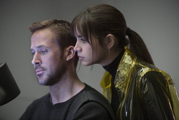 A cinematic still showing two people in profile - a man with short hair and stubble in tactical gear, and a woman with dark hair in a ponytail wearing a yellow protective suit.