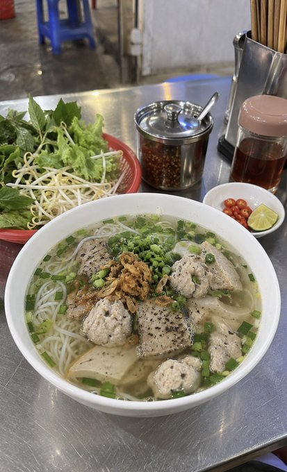 A large bowl of Vietnamese noodle soup topped with meatballs, fried shallots, and green onions, accompanied by a basket of fresh herbs and bean sprouts, chili sauce, and a small dish of lime and chili on a metal table.