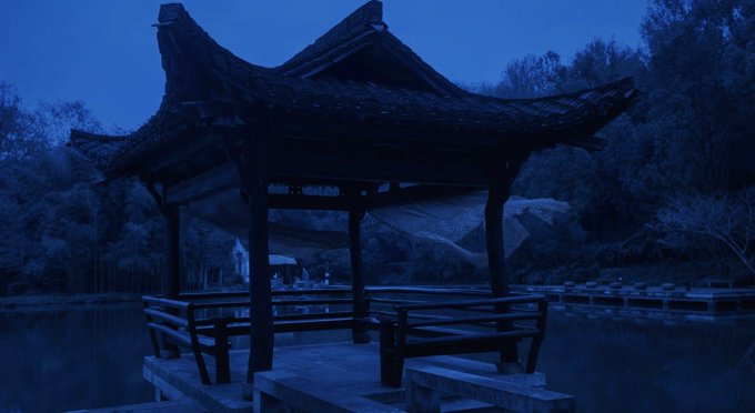 A serene, blue-tinted photograph of a traditional wooden pavilion (gazebo) with a curved roof situated on a platform over water, surrounded by a dark forest setting.