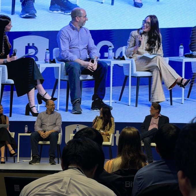 A panel discussion takes place on a stage featuring a large screen displaying a "Bitcoin" logo, where speakers sit in white chairs facing an audience.