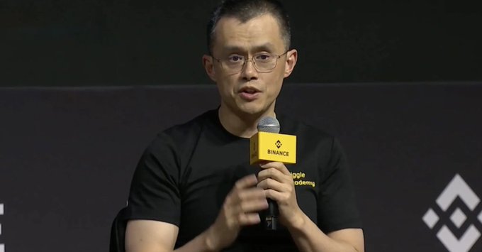 A man with glasses speaks into a microphone bearing the "BINANCE" logo while wearing a black "Binance Academy" t-shirt.