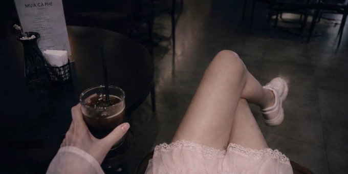 A moody café photo showing a first-person view of a hand holding an iced coffee drink, with crossed legs wearing a light pink dress and sneakers, and a menu reading 'MÙA CÀ PHÊ' on the dark table.