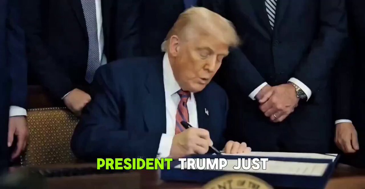 The image shows a man signing a document while wearing a suit and tie, with the text "President Trump Just" written above his signature. The scene takes place at a desk or table where several people are present, including one person sitting on a chair behind the man signing the document. The background of the image is blurred, but it appears to be a room filled with chairs and other people.