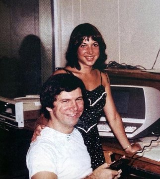 An old photograph shows a smiling man in a white polo shirt seated in front of a woman in a black and white polka-dot dress, with a vintage computer monitor visible behind them.
