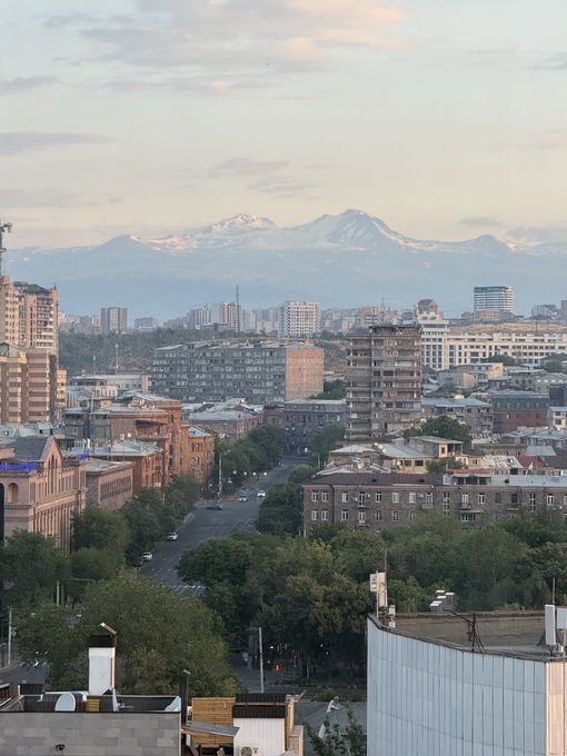 An aerial cityscape view at dusk showing buildings, trees, and streets with snow-capped mountains visible on the horizon under a soft cloudy sky.