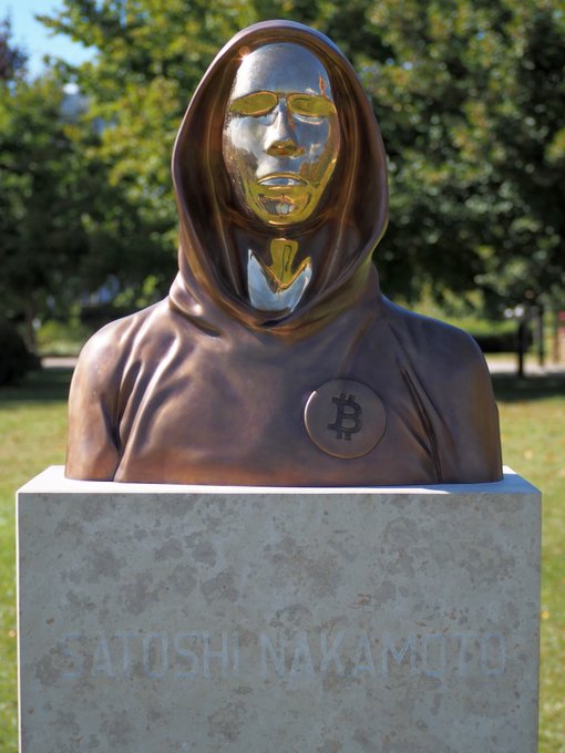 The image features a bronze statue of a man wearing a hooded robe with a face mask, standing on a pedestal. The statue has a "Satoshi Nakamoto" plaque at its base, indicating that it was created by Satoshi Nakamoto, the creator of Bitcoin. The statue is located in an outdoor setting surrounded by trees and grass, suggesting that it might be situated in a park or garden area.