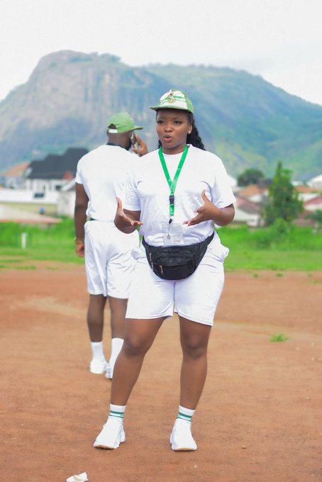 A woman in a white t-shirt, white shorts, and a green cap stands on a dirt field, gesturing while speaking, with another person and a large rocky hill in the background.