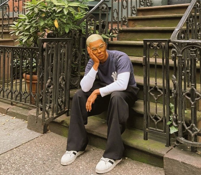 Portrait of a person with short blonde hair wearing glasses and an Adidas t-shirt sitting on stone steps in front of an ornate iron railing.