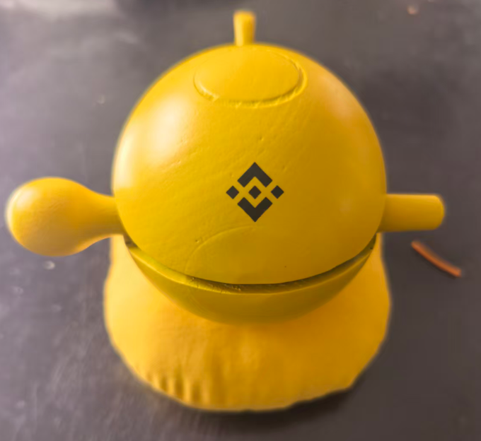 A 3D-printed yellow BMO figure from Adventure Time featuring the Binance logo on its face.