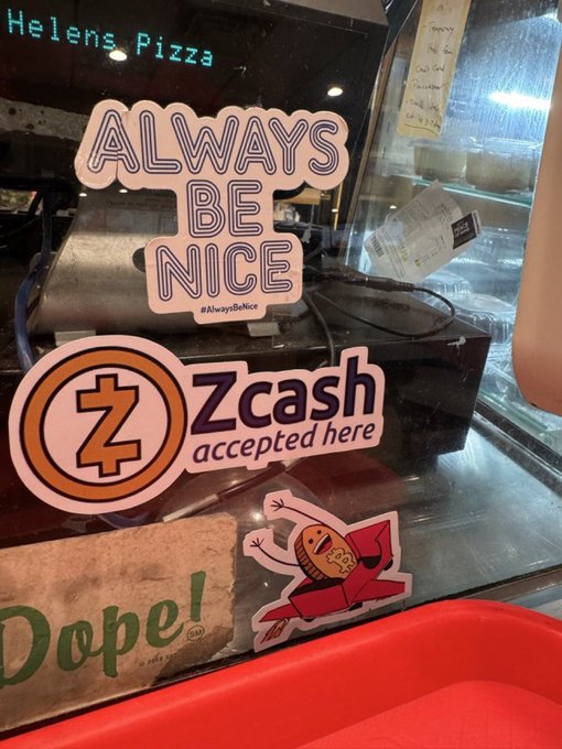 The image features a window display of Helen's Pizza with various stickers and magnets attached to it. One of the stickers reads "Always be nice" while another sticker says "Zcash accepted here". The window display also includes a red tray, which is placed in front of the pizza shop.