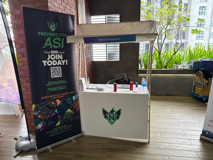 A promotional booth setup for 'NEAR Protocol' and 'ASI' featuring a roll-up banner reading 'PREPARE FOR ASI' and 'Find NEARvana JOIN TODAY!' alongside a white counter with the NEAR logo in what appears to be an event venue.