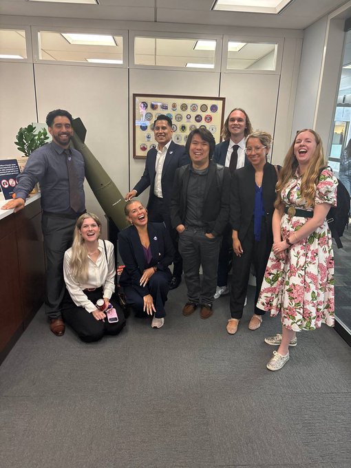 A group photo of eight smiling professionals in business attire posing together in an office setting with a framed emblem display on the wall behind them.
