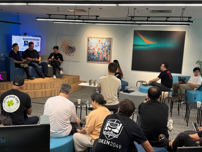 Several people are gathered in a room listening to a panel discussion taking place on a small stage.