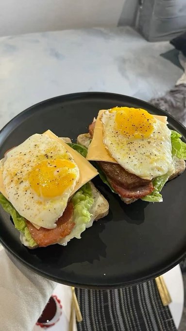 A black plate holds two open-faced breakfast sandwiches featuring sunny-side-up eggs, cheese, bacon, and lettuce on toast.