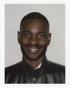 A portrait photograph of a smiling Black man with a short beard, wearing a black jacket with a textured collar.