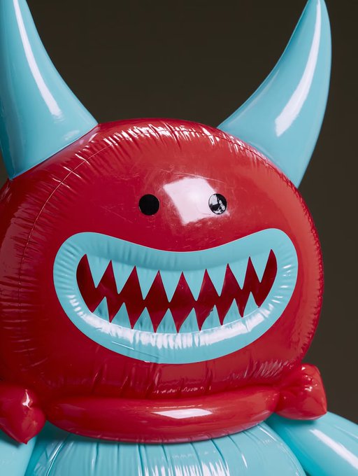 A close-up of a red and light blue inflatable toy figure with horns, a wide jagged smile, and googly eyes against a dark background.