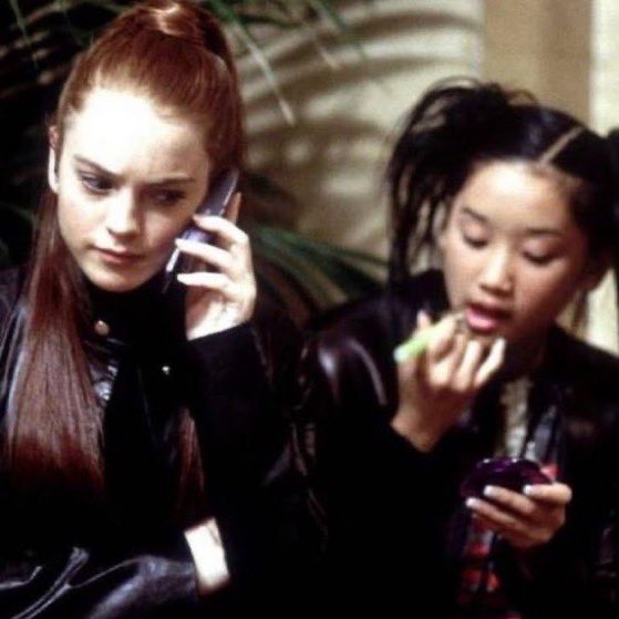 Two young women, one with red hair holding a cell phone and the other applying lipstick while looking in a compact mirror.