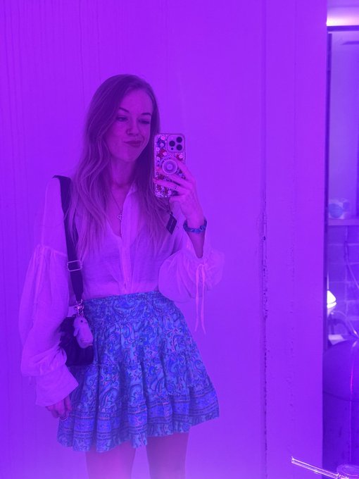 A selfie of a young woman in a white blouse and patterned blue skirt standing in a purple-lit room, taking a photo with a smartphone while carrying a small crossbody bag.
